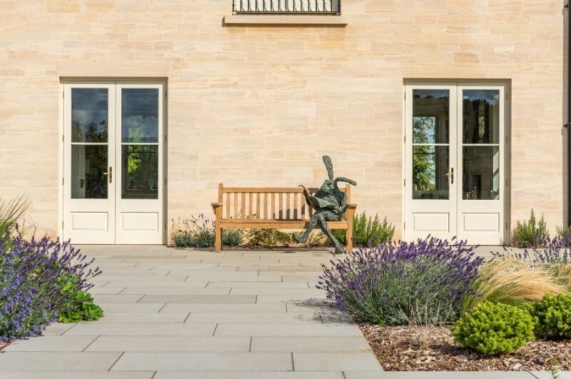 A light stone house with a contrasting grey patio, filled with flowers and a wooden bench in the centre.