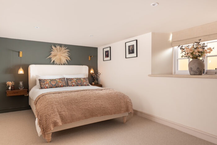 Cosy, modern bedroom with a double bed in the centre of the room and pendant lighting on either side of the bed. A stone windowsill can be seen, dressed with a vase filled with dried flowers.